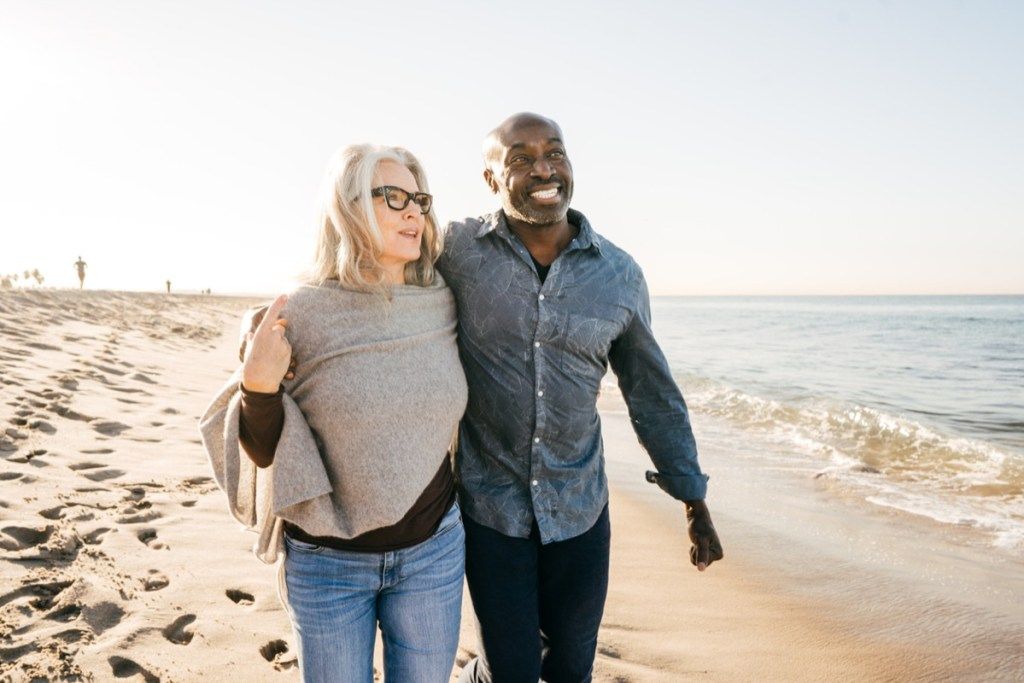 по-възрастна бяла жена двойка чернокож мъж, разхождащ се на плажа с прегърнати ръце