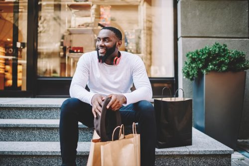   Nasmiješeni muški kupac u trendi odjeći sjedi na stepenicama trgovine s vrećicama s prostorom za kopiranje za etiketu, veseli tamnoputi hipster tip rekreira se nakon kupnje i kupovine