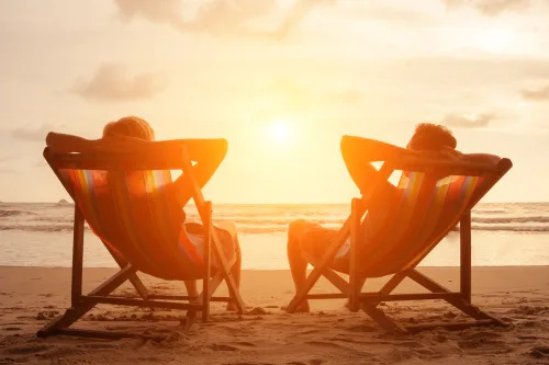   Silhueta srečnega mladega upokojenega para, ki sedi v ležalnikih na plaži ob sončnem zahodu