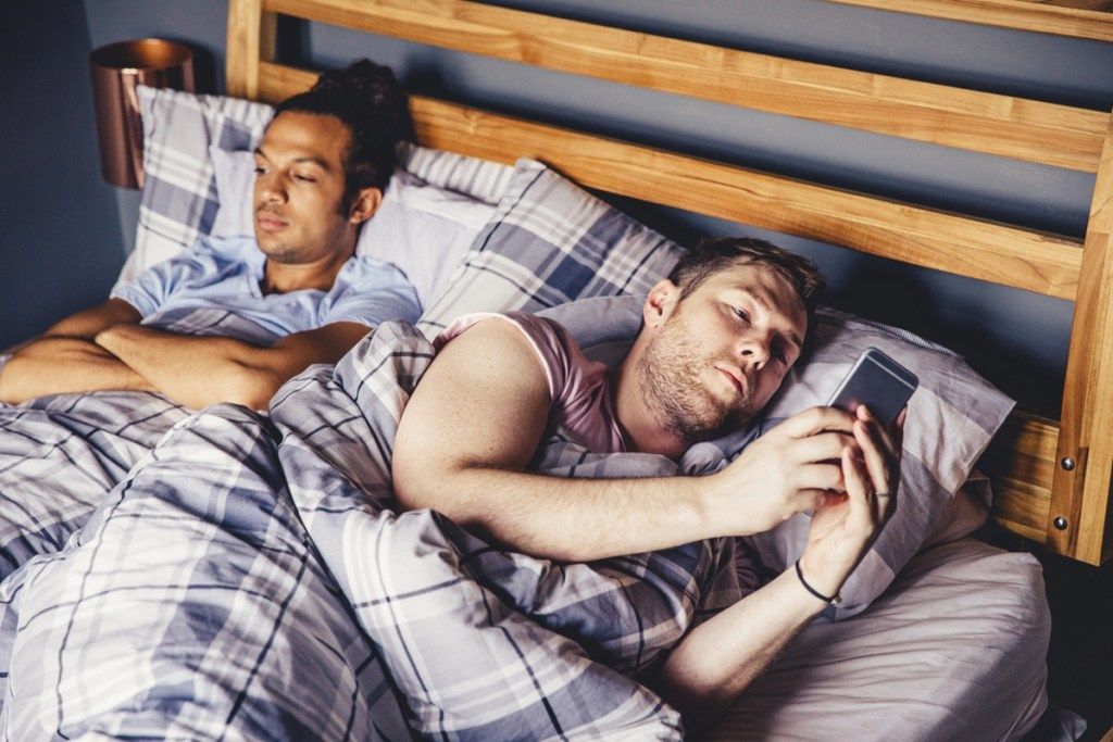Mężczyzna para leży w łóżku rano. Jeden leży na boku i używa swojego smartfona. Drugi ma założone ręce i wygląda, jakby miał dość swojego partnera.