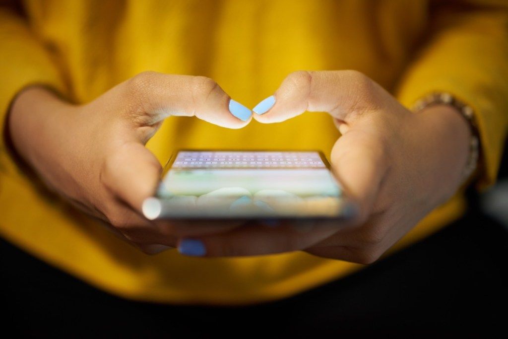 Wanita muda menggunakan telefon bimbit untuk menghantar pesanan teks di rangkaian sosial pada waktu malam. Tutup tangan dengan komputer riba di latar belakang