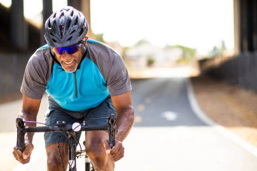   Възрастен чернокож мъж спринтира на своя шосеен велосипед, тренирайки за състезание. Той's smiling and wearing a helmet.