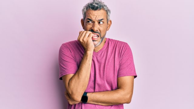   Мъж на средна възраст със сива коса и лилава тениска нервно гризе ноктите си и изглежда несигурен на светло лилав фон