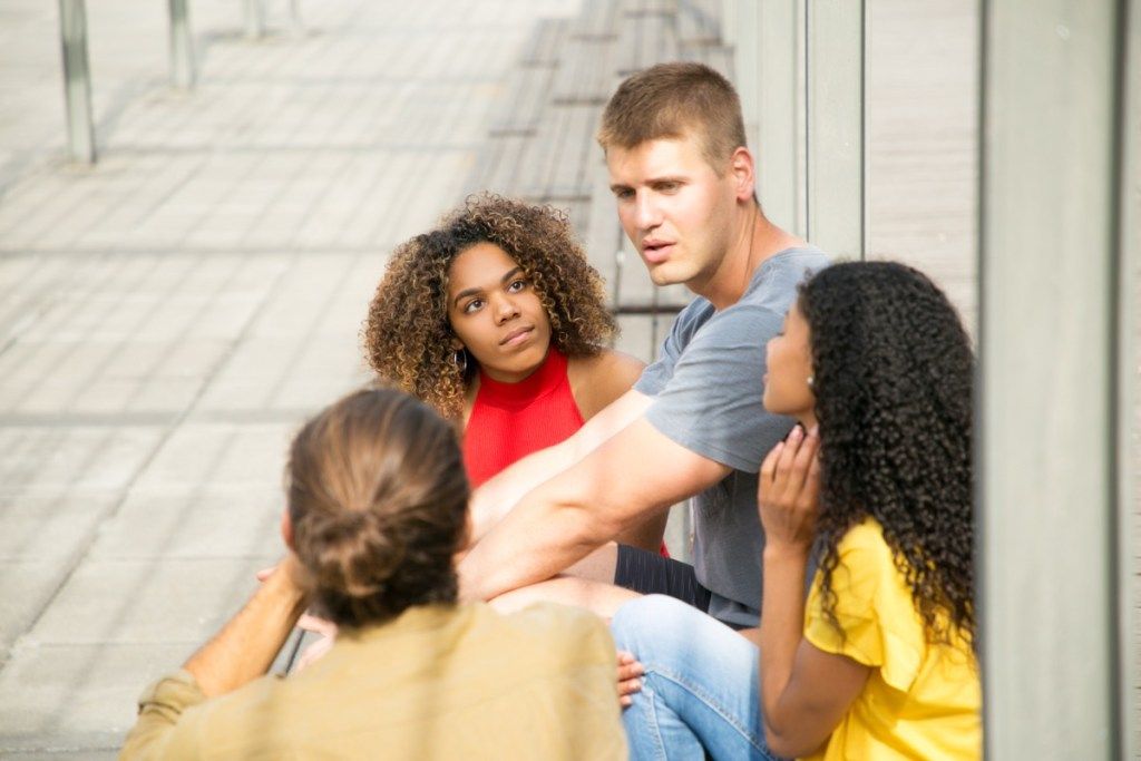 Kaukasier und gemischte Mädchen treffen sich im Freien, um sich zu unterhalten. Interracial Gruppe von jungen Leuten, die auf Außentreppe sitzen, sprechen, zuhören, gestikulieren. Freunde treffen Konzept