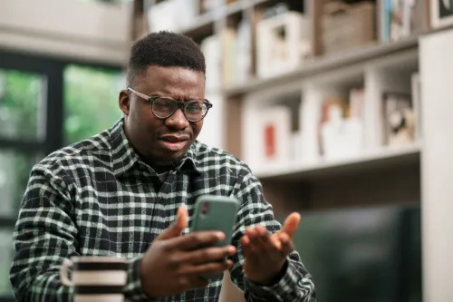   Раздразнен млад мъж седи вкъщи, чете лоши новини на своя смарт телефон с помощта на мобилно приложение и изразява недоволството си