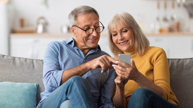   По-възрастна двойка седи на дивана и гледа заедно мобилен телефон.