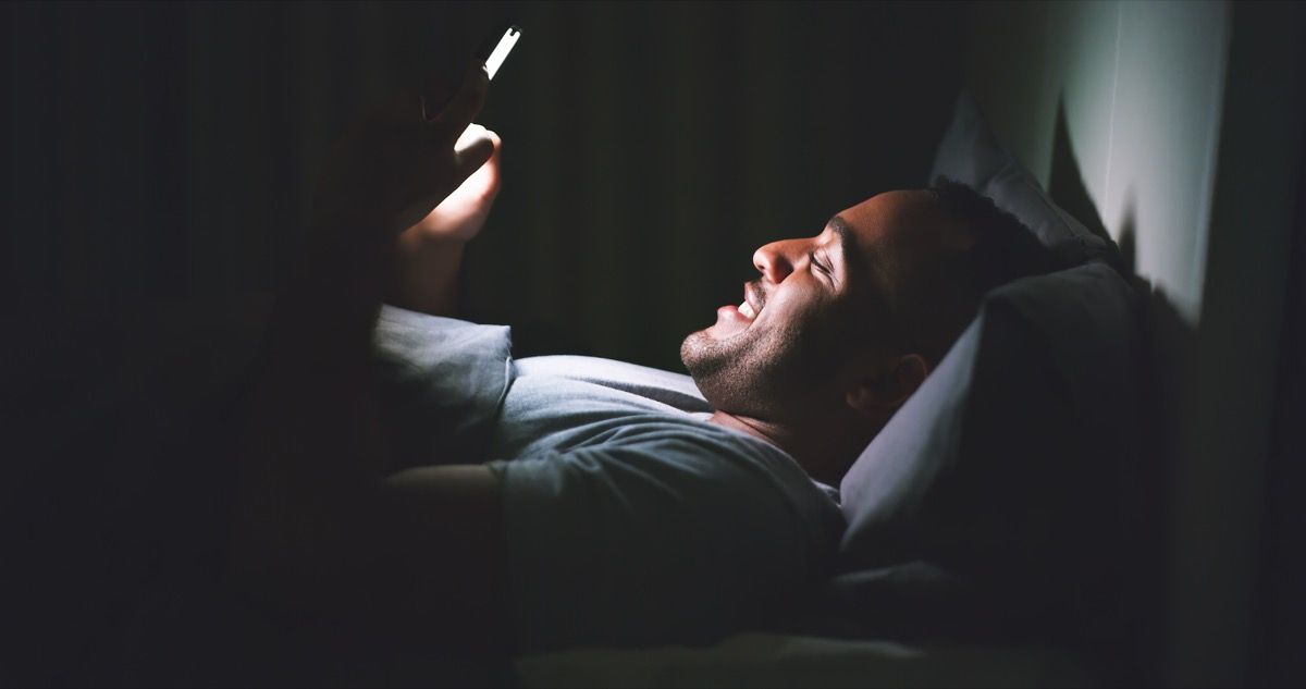 Laukaus iloisesta nuoresta miehestä, joka käyttää matkapuhelinta makaamassa sängyssä myöhään yöllä