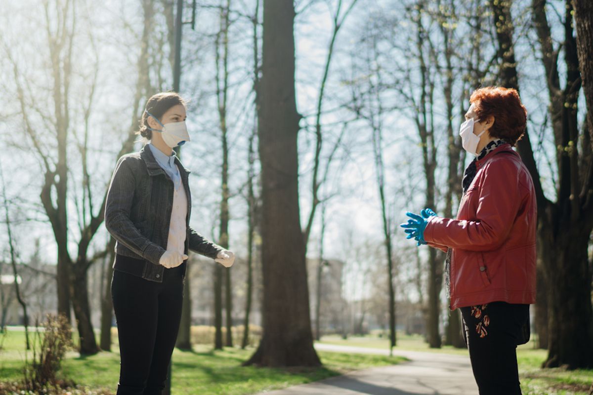 Äldre kvinna med skyddande ansiktsmask / handskar som pratar med en vän utanför