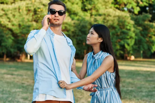   زوجان يتجادلان في حديقة. الرجل يرتدي النظارات الشمسية على الهاتف ولا ينتبه إلى صديقته التي's tugging at his shirt.