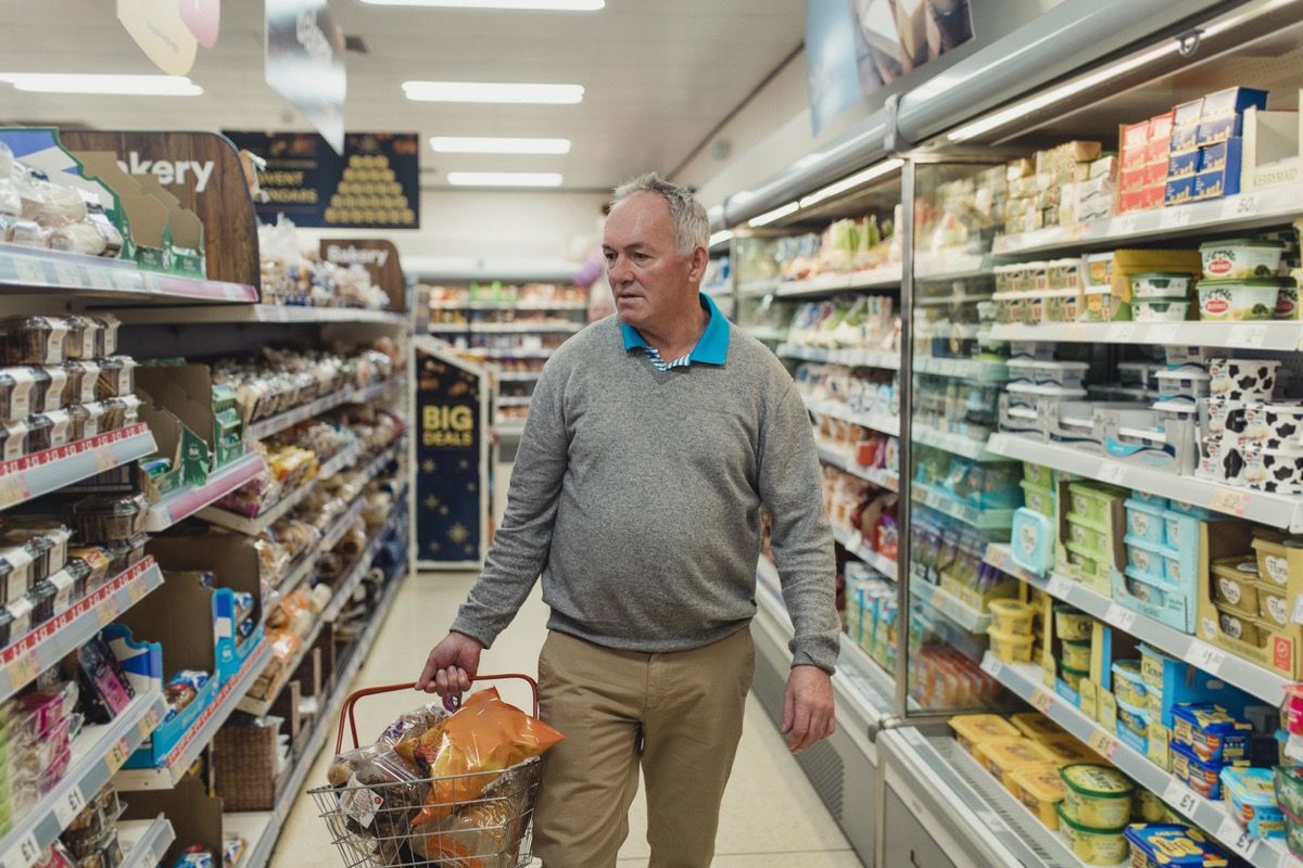 Anciano blanco sosteniendo una canasta y mirando la comida mientras deambula por el pasillo de una tienda de comestibles