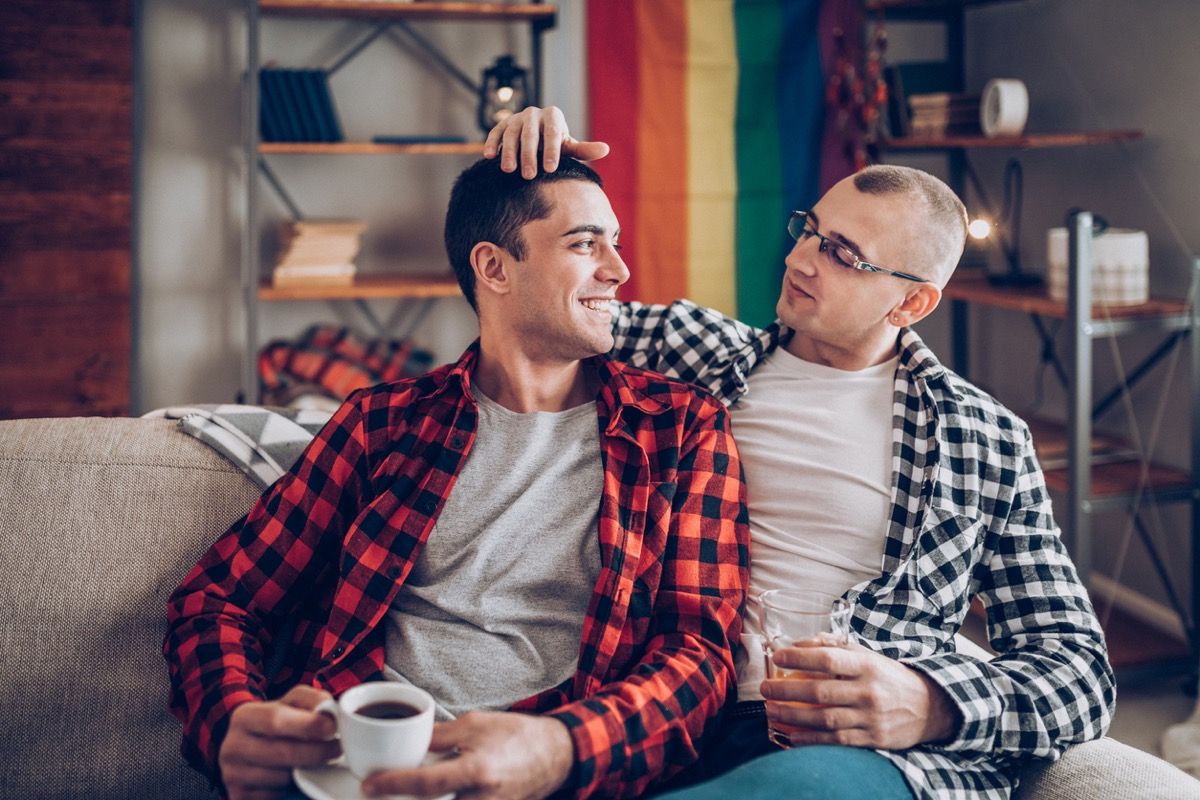 Hombres blancos abrazados en el sofá mientras beben café y agua en su apartamento