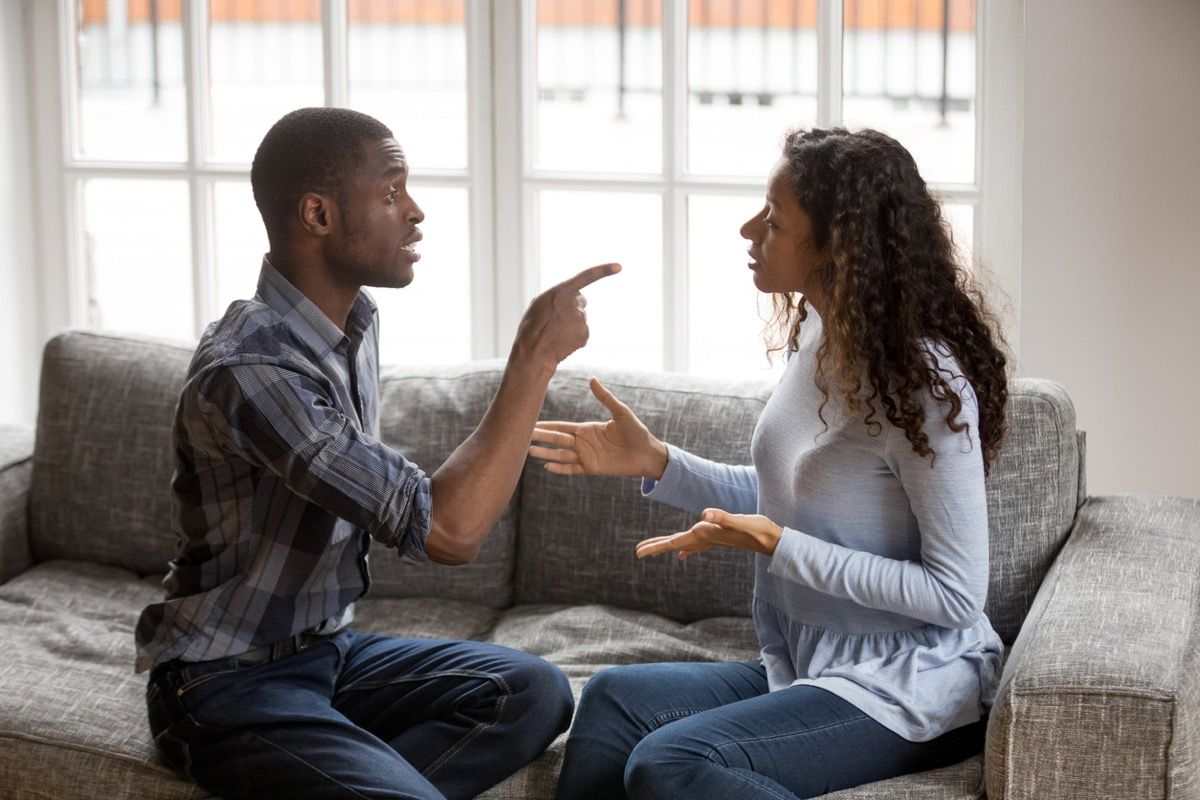 jauns, melns vīrietis, kas ar pirkstu rāda melnajai sievietei, izliekot viņas rokas, kamēr viņi strīdas uz dīvāna