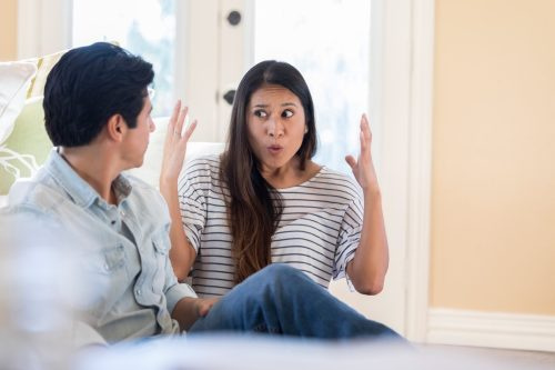   Una mujer adulta de mediana edad levanta las manos con frustración mientras habla con su marido irreconocible en casa. Están sentados en el piso de su sala de estar.