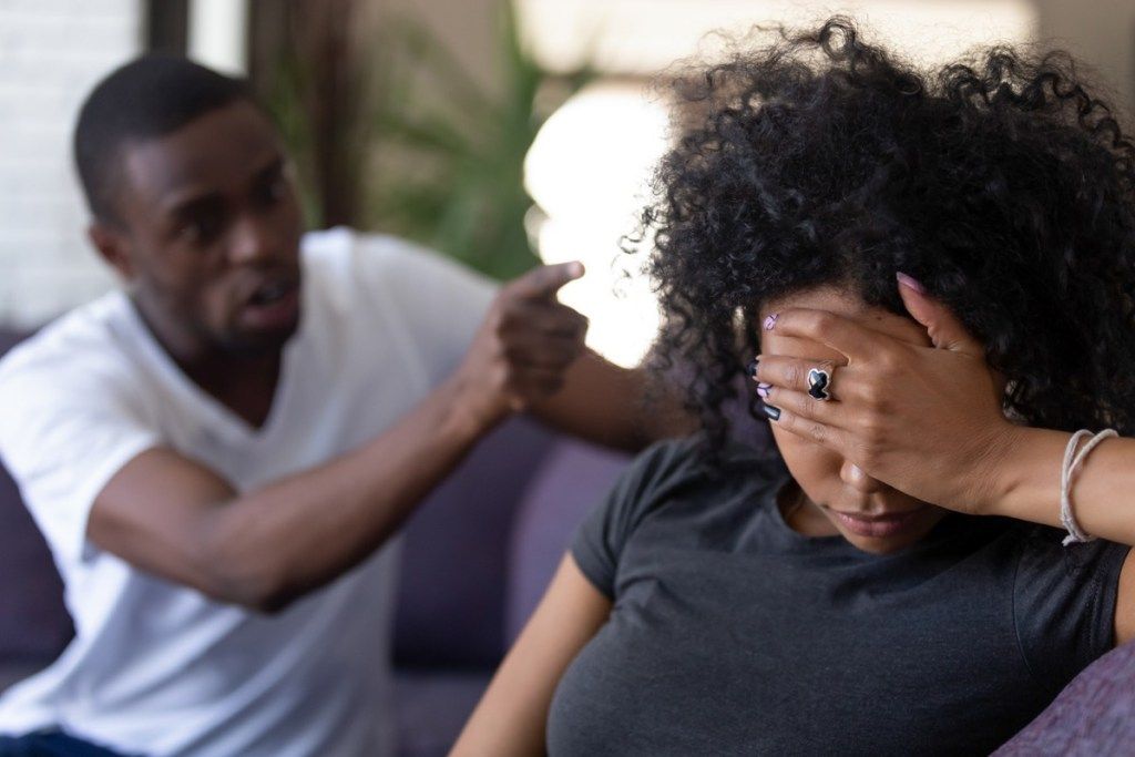 Wanita kulit hitam yang lelah dan frustrasi mengabaikan suaminya yang marah yang menunjukkan jarinya ke arahnya sementara dia menutupi wajahnya di sofa