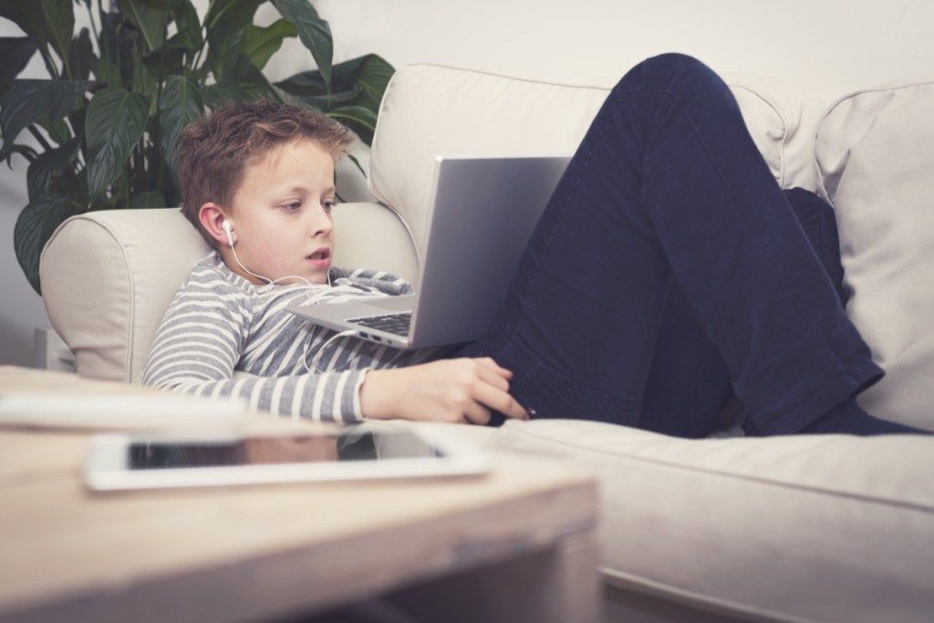 jaunas baltas berniukas ant sofos naudoja nešiojamąjį kompiuterį