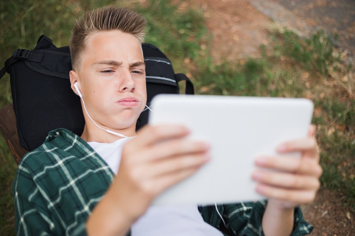 Muchacho adolescente blanco mirando molesto en un ipad mientras está acostado en su mochila afuera