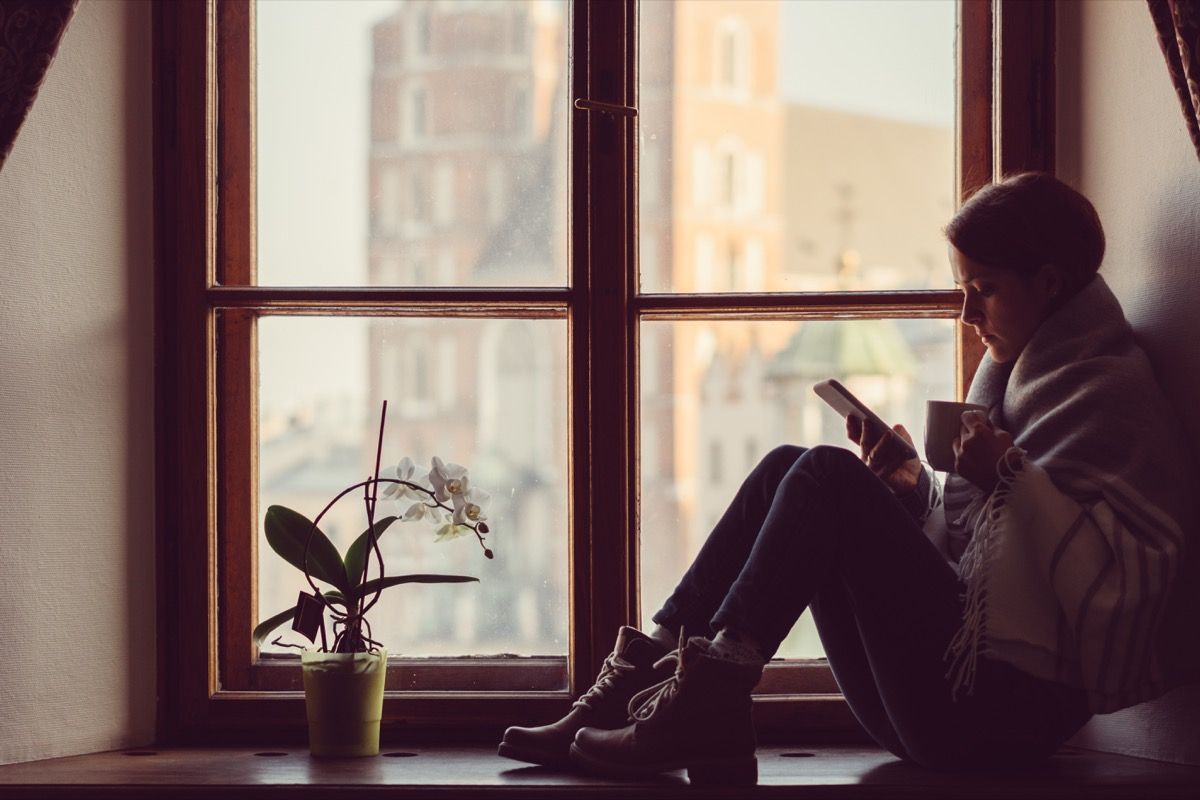 Ženska na oknu, ki pije kavo in brska po mreži na pametnem telefonu
