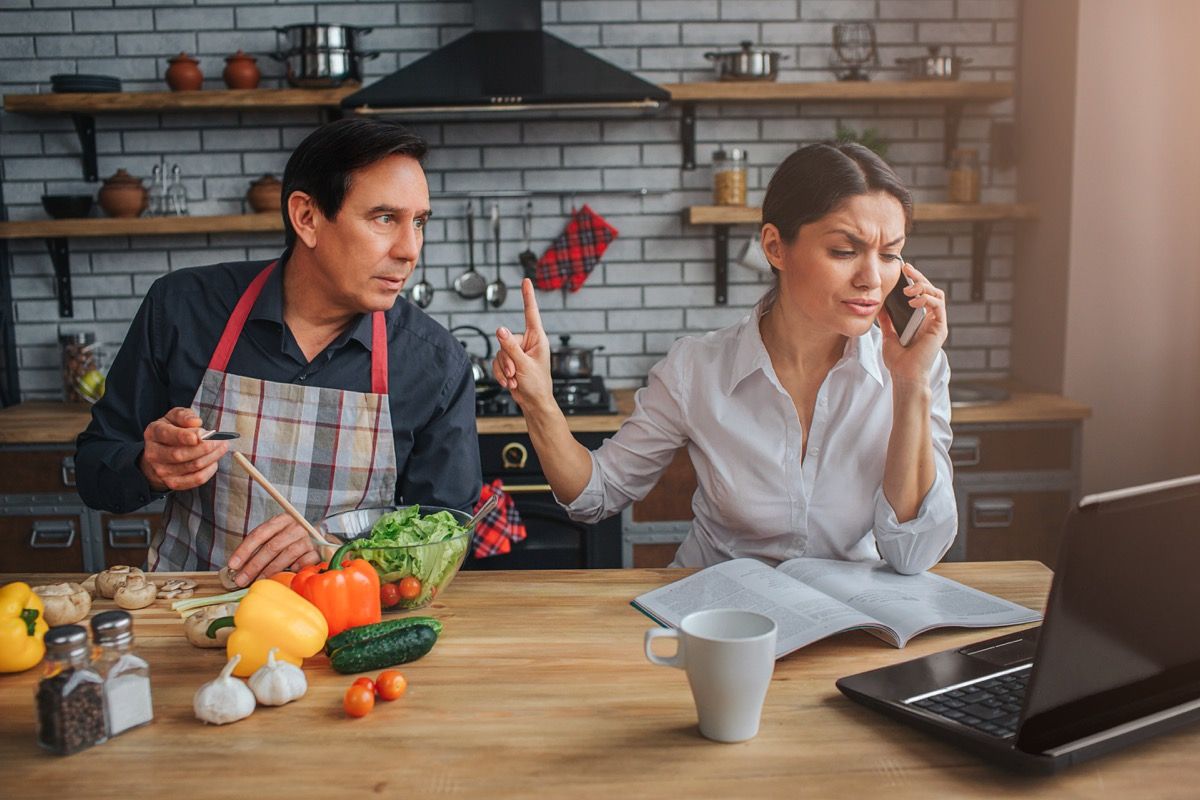 Hombre latino con delantal interrumpe a su esposa en una llamada telefónica en la cocina, etiqueta de más de 40