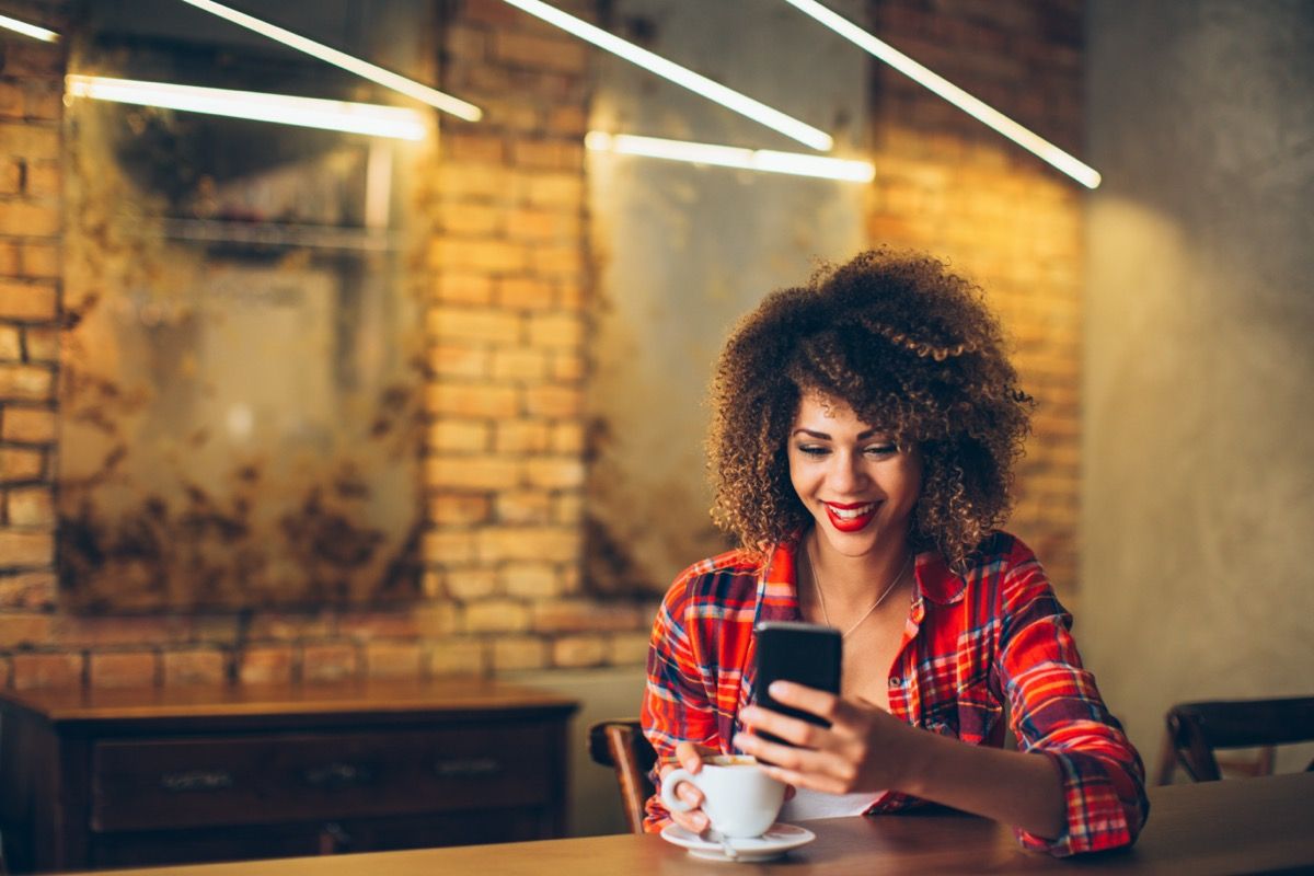 jauna juoda moteris su languotais marškiniais, geria kavą ir šypsosi savo mobiliajam telefonui