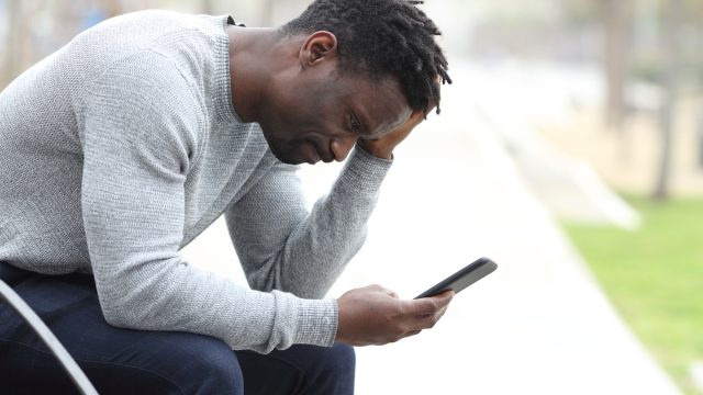   Портрет отстрани на тъжен черен мъж, който проверява мобилния си телефон, докато седи на пейка в парк