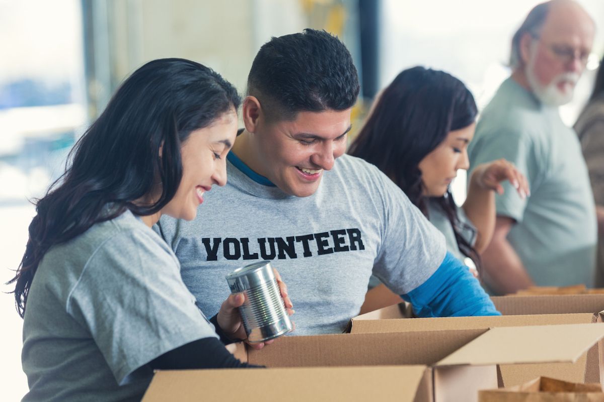 Muškarac i žena Latinx dobrovoljno volontiraju u zajedničkoj banci hrane. Par razvrstava donacije hrane u kartonske kutije, tako da se kutije mogu podijeliti manje sretnim ljudima. Nasmiješeni su i uživaju vraćajući zajednicu. Muškarac nosi majicu VOLONTER.
