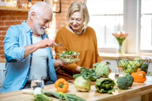   Veseli stariji par jede salatu stojeći zajedno sa zdravom hranom u kuhinji kod kuće