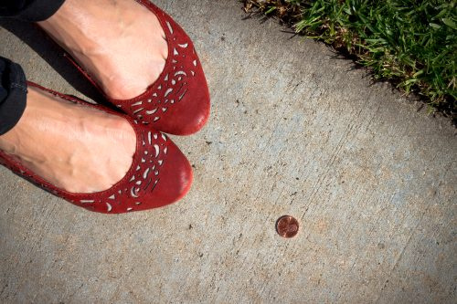   फुटपाथ पर अपने पैरों के पास भाग्यशाली सिर-अप पेनी के साथ लाल फ्लैट जूते में एक महिला का पास से चित्र।