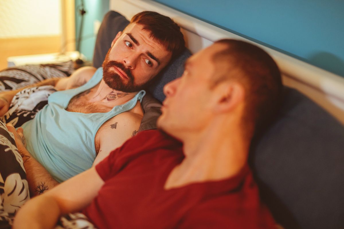 Liūdnas vyras žiūri į šalia lovoje gulintį savo vaikiną