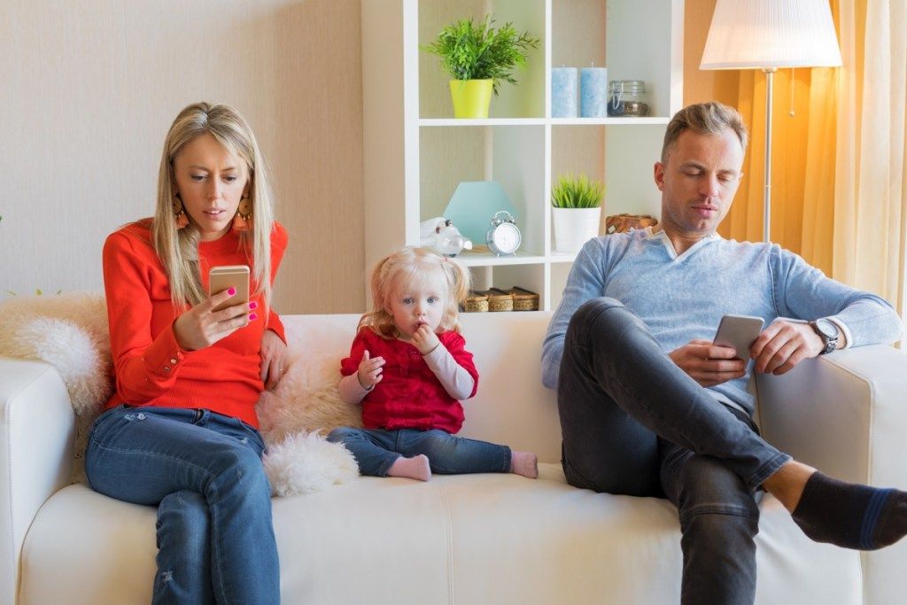 mamma og pappa som sitter på en hvit sofa og leker med mobiltelefoner foran sin lille datter, forbereder barn på skilsmisse