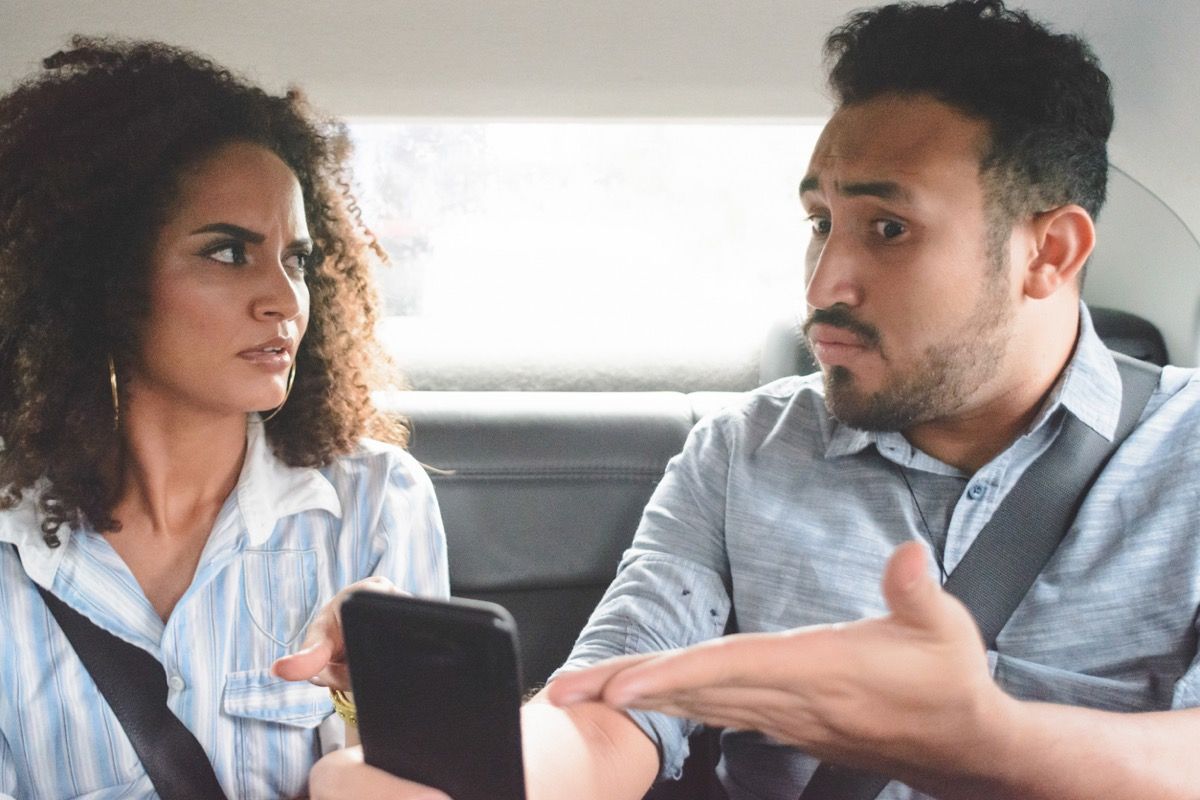 junges Paar streitet auf dem Rücksitz eines Autos, während der Mann defensiv auf sein Handy zeigt
