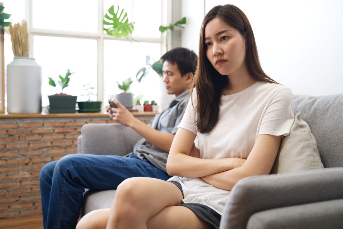 млада двойка, седнала на диван, отделно една от друга, момиче, което изглежда разстроено и мъж по телефона