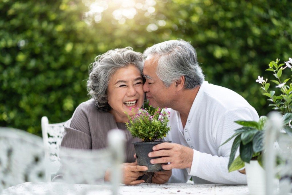 vyresnis azijietiškas vyras, bučiuodamas moterį į skruostą, laikydamas augalą, gailisi per 50