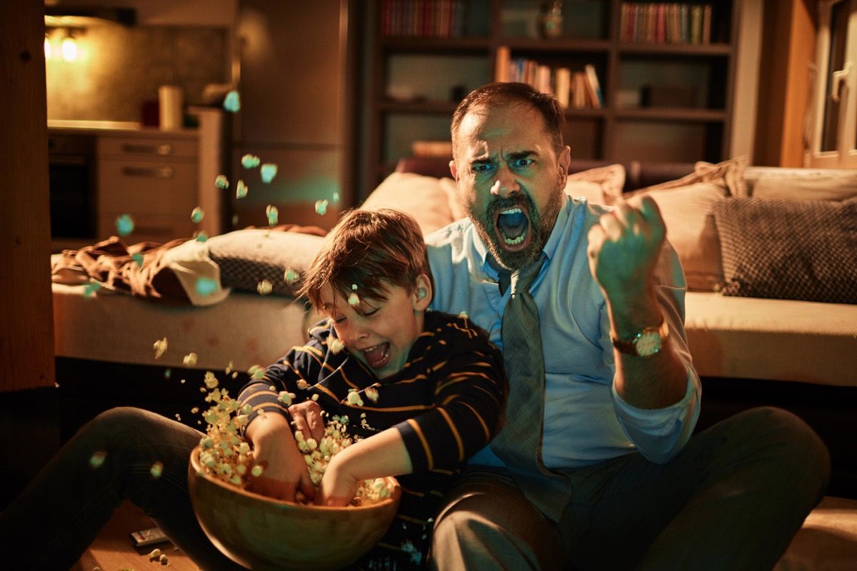 Mann verkleidet sich aufgeregt beim Fernsehen mit Sohn, der Popcorn wirft