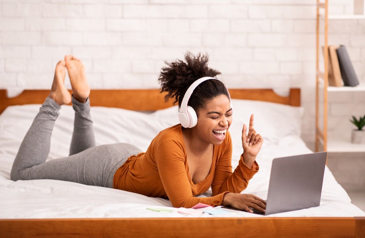 बिस्तर पर युवा अश्वेत महिला अपने कंप्यूटर पर हेडफोन के साथ गाती हुई