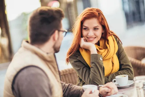   Двойка, седнала в кафене на открито; жената се усмихва и се взира в партньора си.