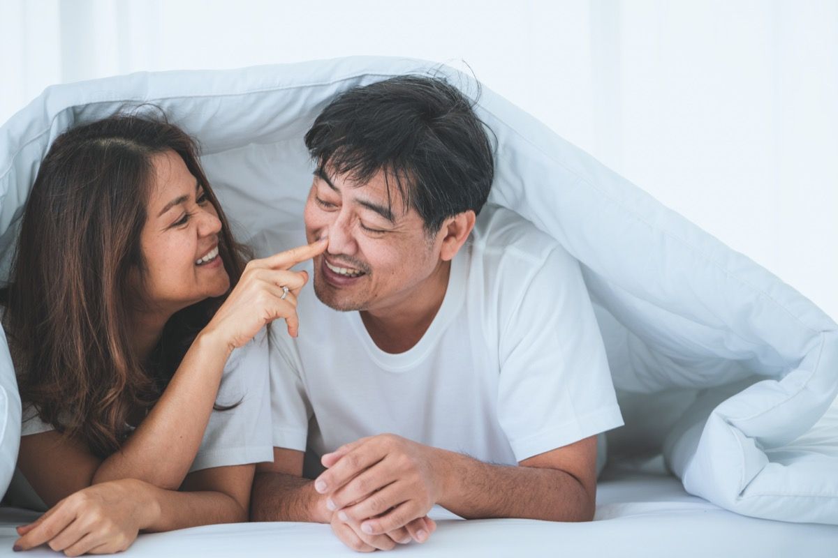 vanhempi aasialainen nainen hymyilee ja tönäisee vanhemman aasialaisen miehen nenää valkoisen lohduttajan alla