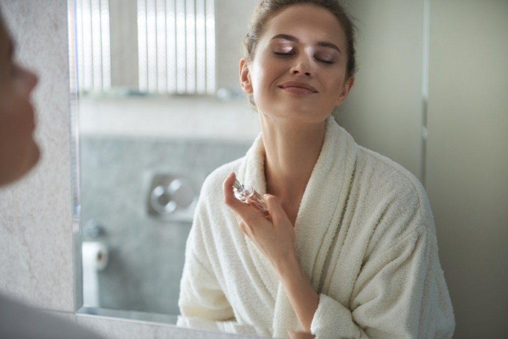 Conceptul de îngrijire personală. Reflectarea în talie a unei tinere zâmbitoare în halat de baie alb, bucurându-se de dimineața în timp ce aplică parfumul preferat în fața oglinzii din baie (Concept de îngrijire personală.