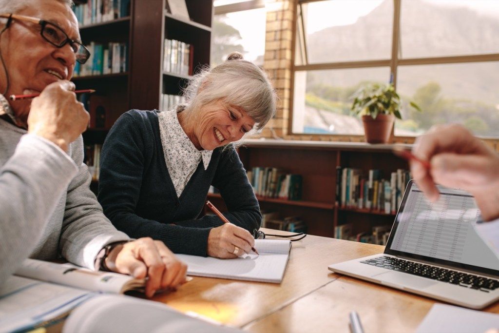 Vanem naine, kes kirjutab märkmeid, istub raamatukogus koos oma meeskolleegidega. Eakad inimesed ülikoolis arutlevad ja õpivad.