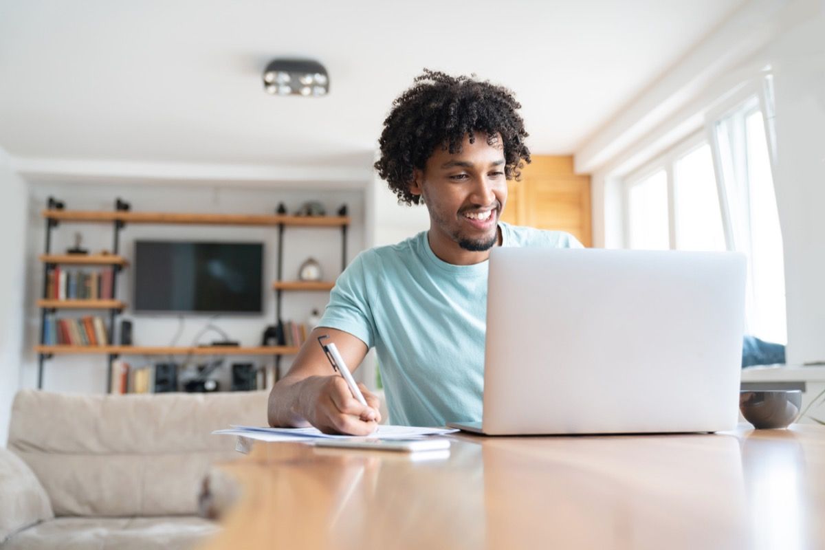 Joven negro sonriendo mientras escribe algo y mira su computadora portátil