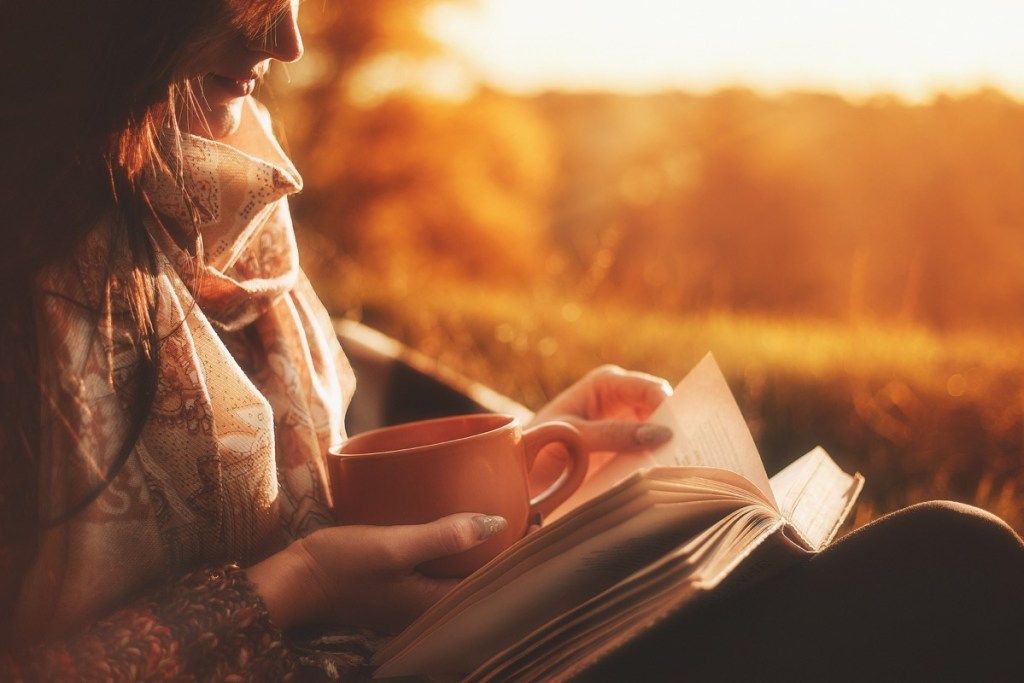 امرأة تقرأ كتابًا في الخارج عند غروب الشمس تحمل فنجانًا من القهوة - كتب كلاسيكية
