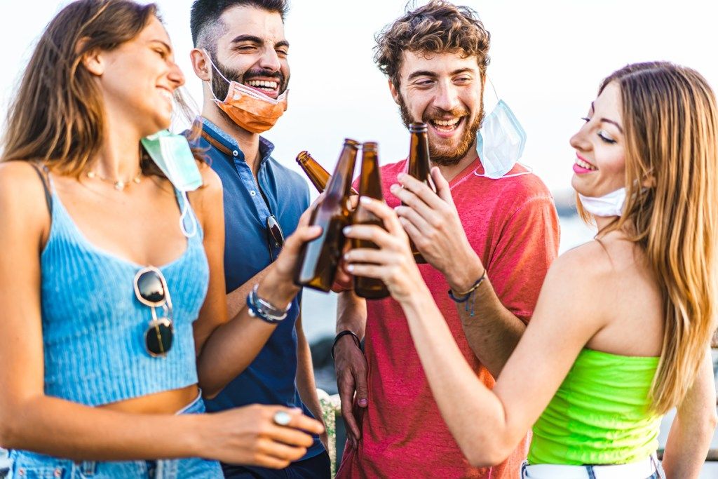 Četru jaunu vīriešu un sieviešu grupa uzmundrina alus pudeles kopā ar sejas maskām, kas atvieglo koronavīrusa izplatīšanos