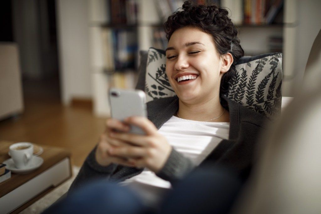فون پر رہتے ہوئے ہنستے ہوئے گھر میں آرام سے ہفتے کے آخر میں لطف اٹھانے والی نوجوان عورت