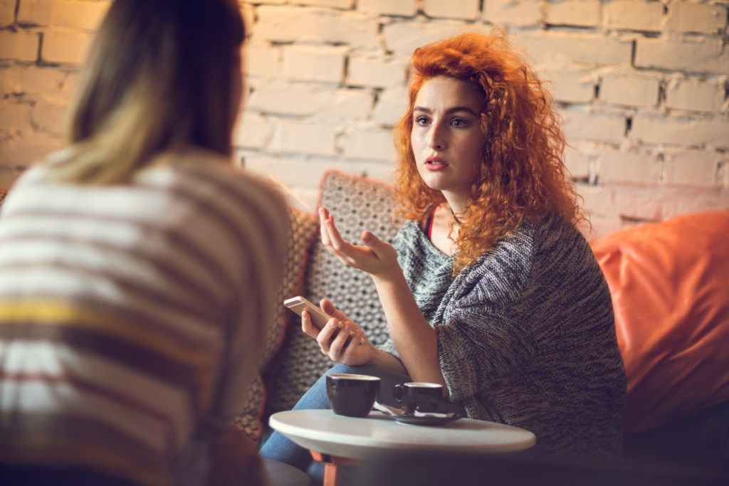 jauna raudonplaukė moteris, sėdinti kavinėje su drauge ir kalbanti apie kažką.