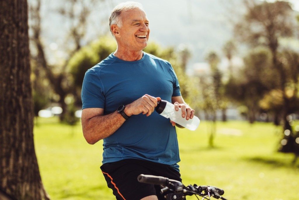 Старши мъж във фитнес носи питейна вода, седнал на велосипеда си. Весело лице за фитнес, което прави почивка по време на колоездене в парк.