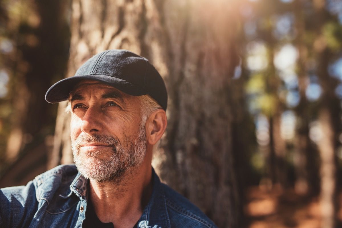 starejši bel moški v črnem klobuku, ki sedi pred velikanskim drevesom in je videti miren