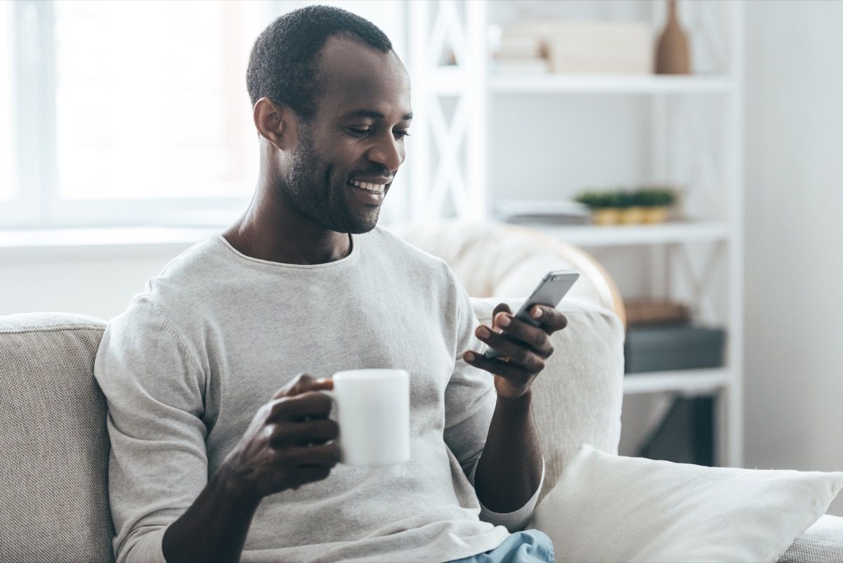 Hombre negro de mediana edad enviando mensajes de texto en el sofá con una taza de té