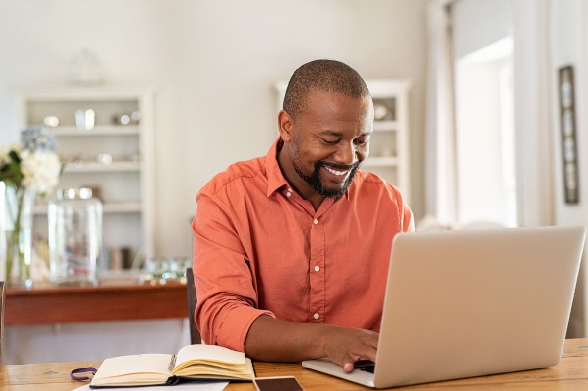 लिविंग रूम में घर पर लैपटॉप का उपयोग करते हुए मुस्कुराते हुए काले आदमी। खुश परिपक्व व्यापारी ईमेल भेजें और घर पर काम कर रहे हैं। अफ्रीकी अमेरिकी फ्रीलांसर टेबल पर पेपरवर्क और दस्तावेजों के साथ कंप्यूटर पर टाइपिंग करता है।