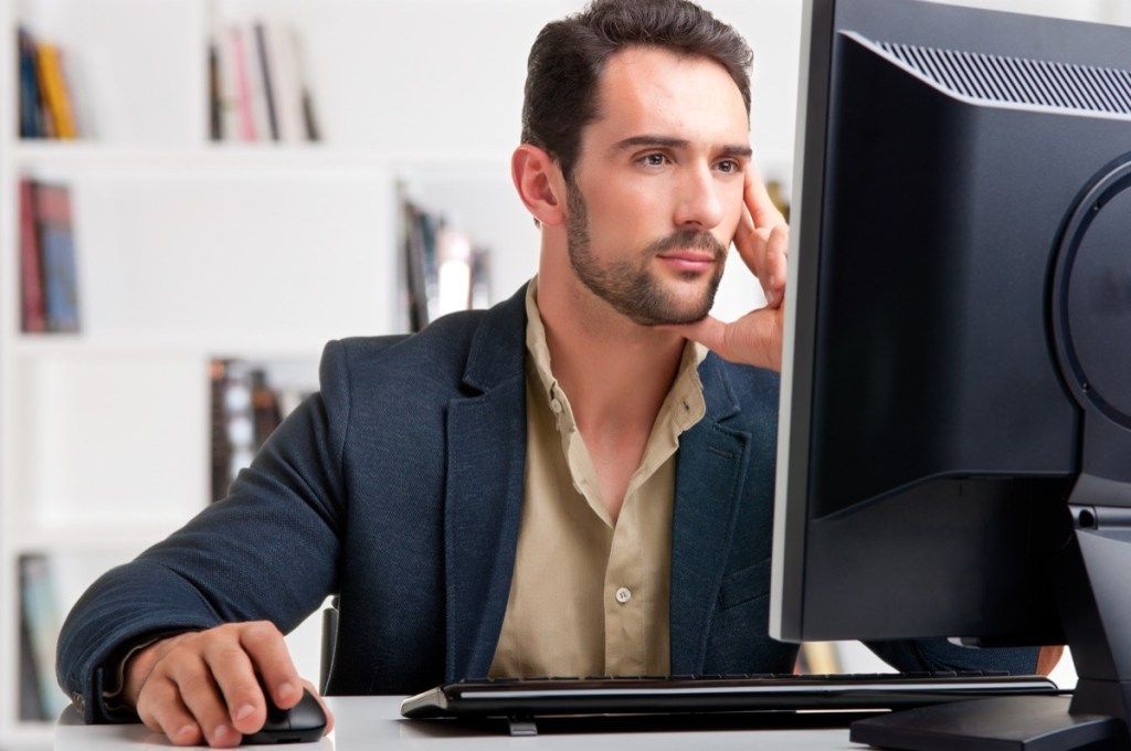 keski-ikäinen valkoinen mies työskentelee suurella tietokonenäytöllä työskennellessään kotona wfh-toimistossa