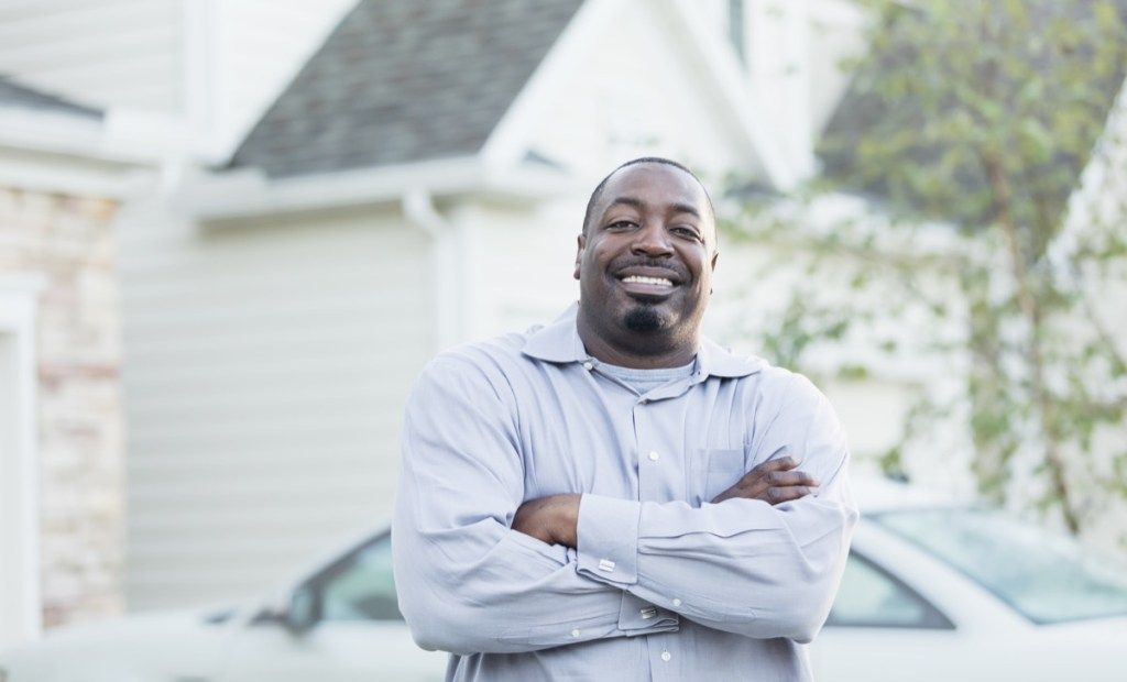 Nobriedis afroamerikāņu vīrietis ap 40 gadiem, kas stāv brīvā dabā pie savas mājas piebraucamā ceļa un smaida kamerai ar sakrustotām rokām.
