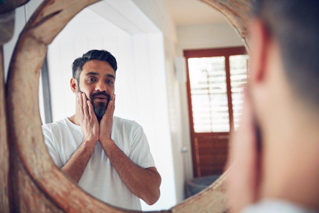 Foto de um homem maduro olhando para seu reflexo no espelho do banheiro